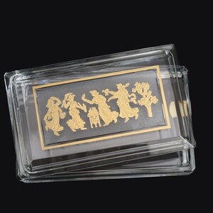 May include: A clear, rectangular box with a sliding lid. Inside, a gold-colored rectangular insert features a relief of figures in a procession. The box is made of clear plastic and sits against a black background.