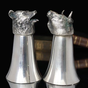 May include: A pair of silver-colored salt and pepper shakers. One shaker is topped with a bear head, the other with a bull head. The shakers have a flared base and are set against a dark background.