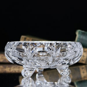 Puede incluir: Un cuenco de cristal tallado transparente, de forma redondeada, con tres pequeños pies decorativos. El cuenco presenta un detallado patrón de hojas y geométrico en el borde. El fondo es oscuro, con libros borrosos visibles.