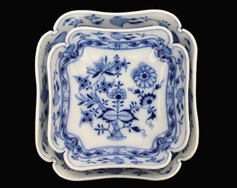 Op de afbeelding: Set van drie witte porseleinen serveerschalen met een blauw bloemmotief. De schalen hebben een vierkante vorm met geschulpte randen. Het blauwe bloemmotief omvat bloemen, bladeren en wijnranken. De schalen zijn genest.
