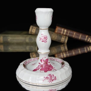 Op de afbeelding: Witte porseleinen kandelaar met roze bloemmotieven en gouden randen. De kandelaar heeft een ronde basis met een geschulpte rand en een centrale stam. Het bloemmotief herhaalt zich op de basis en de stam. De achtergrond is zwart.