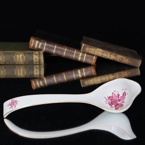 May include: A white porcelain gravy ladle with a pink floral design. The ladle has a long handle and a wide, shallow bowl.
