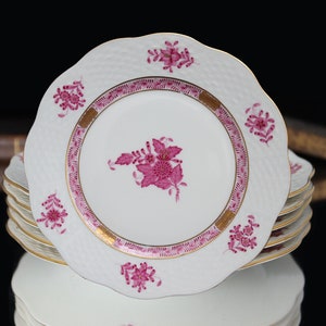 May include: A stack of white porcelain plates with scalloped edges. Each plate features a pink floral design in the center and around the rim, with gold accents. The plates have a textured surface and are stacked on a reflective surface.