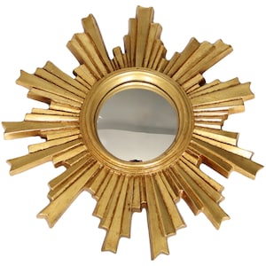 May include: A round, gold-colored mirror with a sunburst design. The mirror has a circular reflective surface surrounded by radiating, textured gold rays. The frame is made of a metallic material.