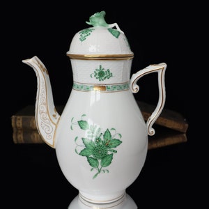 Könnte beinhalten: Eine weiße Porzellan-Kaffeekanne mit grünem und goldenem Blumendesign. Die Kanne hat einen Deckel mit einem grünen Blumenknauf.