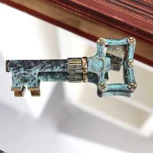 Könnte beinhalten: Ein Schlüssel im Vintage-Stil mit einer grünen Patina und goldenen Akzenten. Der Schlüssel hat ein dekoratives Design auf dem Bogen und ein kleines Loch in der Mitte.
