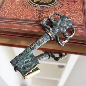 Könnte beinhalten: Ein antiker Schlüssel mit aufwendigem Design und verwitterter, grünspanfarbener Oberfläche mit goldenen Akzenten. Er liegt auf einem Vintage-Buch mit rotem Einband und goldener Schrift, was auf Geschichte oder Literatur hindeutet.