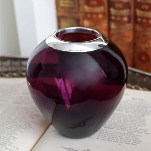 Peut inclure: Un vase rond en verre violet fonc&eacute; avec un bord argent&eacute;. Le vase est pos&eacute; sur un livre ouvert. L'arri&egrave;re-plan comprend une pile de vieux livres.