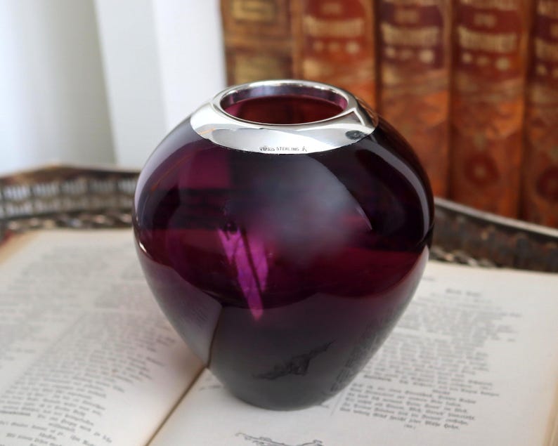 Peut inclure: Un vase rond en verre violet fonc&eacute; avec un bord argent&eacute;. Le vase est pos&eacute; sur un livre ouvert. L'arri&egrave;re-plan comprend une pile de vieux livres.
