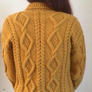 May include: Mustard yellow cable-knit sweater with diamond and braid patterns. The back of the sweater is visible, showcasing the intricate knit design. The sweater has a ribbed collar and cuffs, and is a warm, cozy garment.