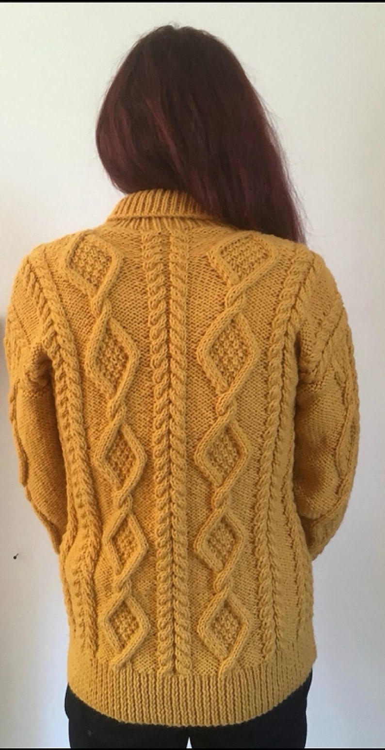 May include: Mustard yellow cable-knit sweater with diamond and braid patterns. The back of the sweater is visible, showcasing the intricate knit design. The sweater has a ribbed collar and cuffs, and is a warm, cozy garment.