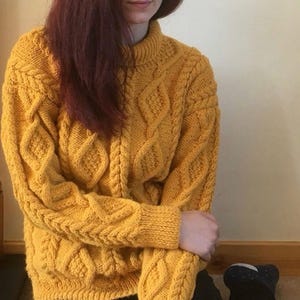 May include: Mustard yellow cable-knit jumper with a high, ribbed collar. The jumper features a textured diamond and braid pattern. The sleeves and cuffs are ribbed. The person is wearing black trousers.