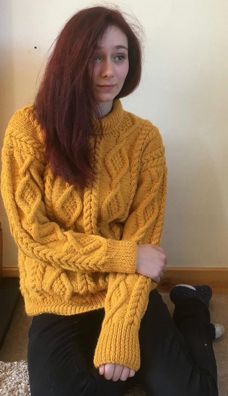 May include: Mustard yellow cable-knit jumper with a high, ribbed collar. The jumper features a textured diamond and braid pattern. The sleeves and cuffs are ribbed. The person is wearing black trousers.