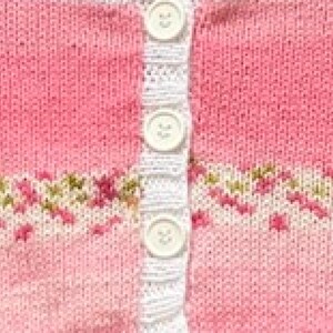 May include: A pink knitted cardigan featuring a vertical white button placket with three white buttons. The garment displays a horizontal band of intricate floral-inspired patterns in shades of green, white, and pink across the mid-section.