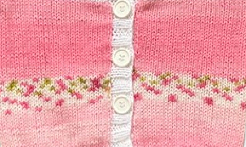 May include: A pink knitted cardigan featuring a vertical white button placket with three white buttons. The garment displays a horizontal band of intricate floral-inspired patterns in shades of green, white, and pink across the mid-section.