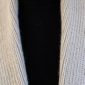 Op de afbeelding: Een beige gebreid vest met een brede sjaalkraag met een geribbelde textuur, gedragen over een effen zwart shirt met een opstaande kraag. Het vest heeft een consistent, dicht breipatroon over de voorpanden en de opvallende kraagstructuur.