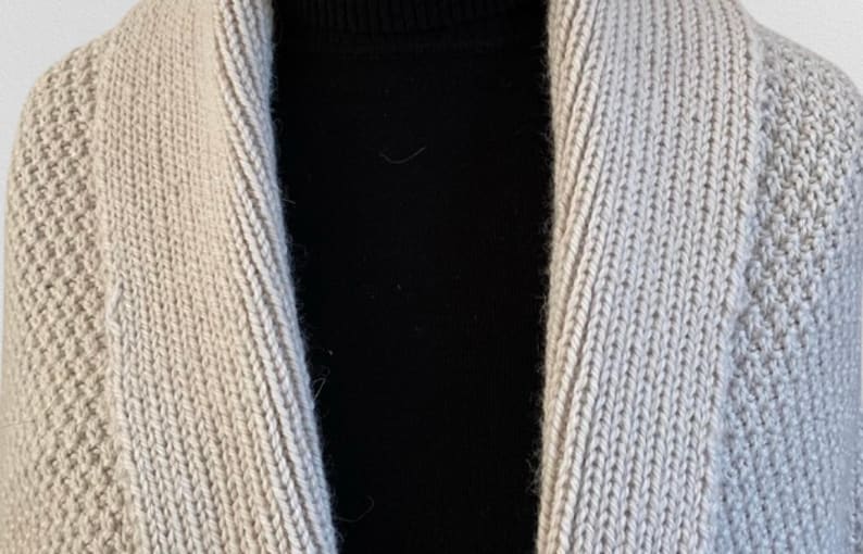 Op de afbeelding: Een beige gebreid vest met een brede sjaalkraag met een geribbelde textuur, gedragen over een effen zwart shirt met een opstaande kraag. Het vest heeft een consistent, dicht breipatroon over de voorpanden en de opvallende kraagstructuur.
