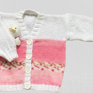 May include: A long-sleeved knit cardigan featuring a white upper section and a pink lower section with a small floral pattern. The garment has a V-neckline, a white button-down front with four white buttons, and ribbed cuffs and hem.