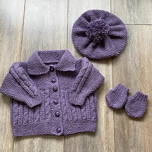 May include: A knitted lavender baby set including a button-up cardigan, a beret with a pom-pom, and a pair of mittens. The set is made of a soft, textured yarn and features a cable knit design on the cardigan.