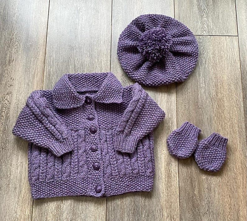 May include: A knitted lavender baby set including a button-up cardigan, a beret with a pom-pom, and a pair of mittens. The set is made of a soft, textured yarn and features a cable knit design on the cardigan.