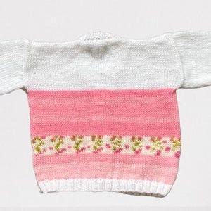 May include: A long-sleeved knit jumper featuring a white upper section and a pink lower section. The lower portion includes a horizontal band with a small floral pattern in shades of green and pink, finished with a white ribbed hem and cuffs.