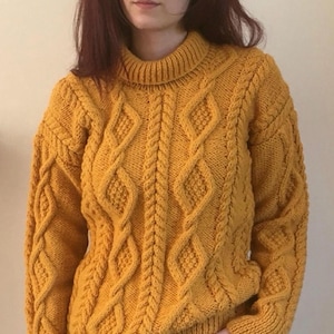May include: Mustard yellow cable-knit jumper with a high neckline. The jumper features a diamond and braid pattern across the front and sleeves. The sleeves have ribbed cuffs. The person is wearing black trousers.