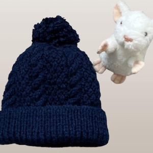 May include: A dark navy blue knit beanie featuring a textured cable knit pattern and a matching pom-pom on top. Beside the beanie is a small, white plush mouse toy with light pink ears and paws, depicted in a standing position with arms raised.