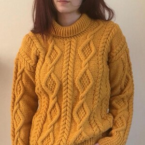 May include: Mustard yellow cable-knit jumper with a high neckline. The jumper features a diamond and braid pattern across the front and sleeves. The sleeves have ribbed cuffs. The person is wearing black trousers.
