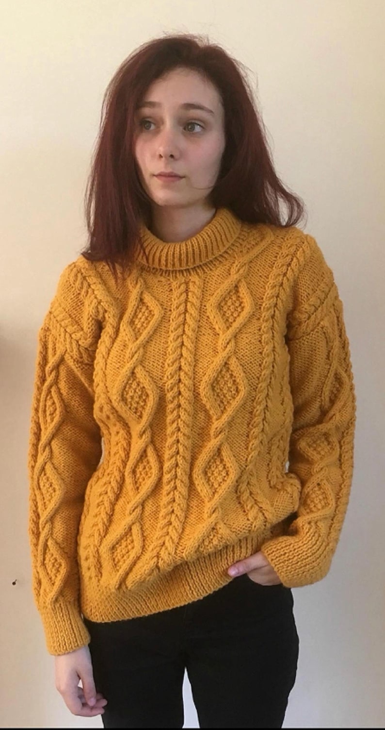 May include: Mustard yellow cable-knit jumper with a high neckline. The jumper features a diamond and braid pattern across the front and sleeves. The sleeves have ribbed cuffs. The person is wearing black trousers.