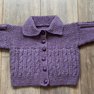 May include: A hand-knitted, lavender-colored cardigan with a collared neckline and button-down front. The sweater features a textured stitch pattern on the body and sleeves, with a cable knit design on the lower front. The sleeves are long and the cardigan is laid flat.