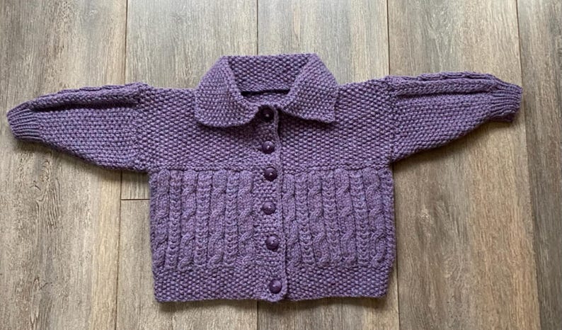 May include: A hand-knitted, lavender-colored cardigan with a collared neckline and button-down front. The sweater features a textured stitch pattern on the body and sleeves, with a cable knit design on the lower front. The sleeves are long and the cardigan is laid flat.
