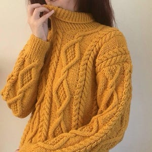 May include: Mustard yellow cable-knit jumper with a roll neck. The jumper features a diamond and braid pattern. The sleeves and body have a textured design. The garment is made of a thick, warm material, perfect for cold weather.