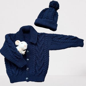 May include: A navy blue cable-knit cardigan with a collar and four buttons, paired with a matching beanie featuring a pom-pom. A small white plush mouse toy is tucked into the sleeve of the cardigan, which features a textured pattern.