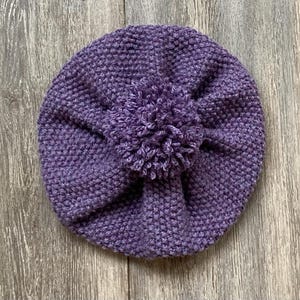 May include: A knitted purple hat with a textured stitch pattern and a large pom-pom on top. The hat is round and appears to be handmade, perfect for keeping warm in cold weather. The hat is shown on a wooden surface.