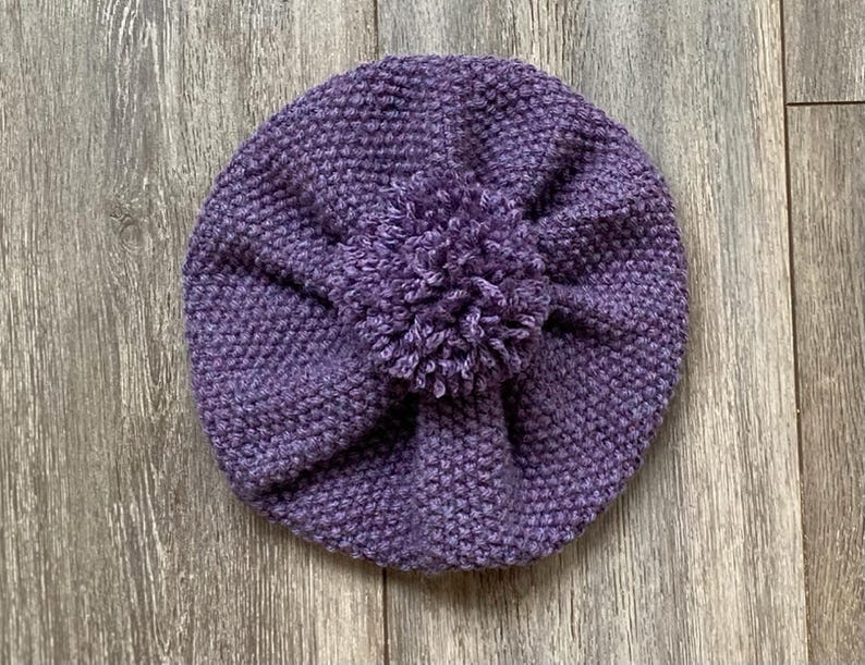 May include: A knitted purple hat with a textured stitch pattern and a large pom-pom on top. The hat is round and appears to be handmade, perfect for keeping warm in cold weather. The hat is shown on a wooden surface.