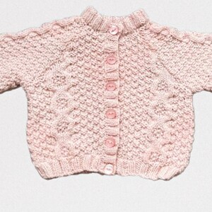 Op de afbeelding: Een lichtroze gebreid babyvestje met een ronde kraag en lange mouwen. De trui heeft een textuurpatroon en wordt gesloten met vijf roze knopen. De mouwen hebben een kabelgebreid ontwerp.