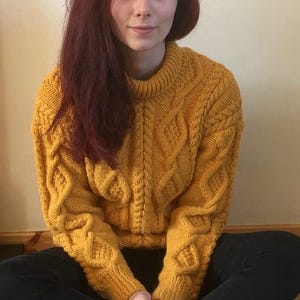 May include: A mustard yellow cable-knit jumper with a crew neck and diamond pattern on the sleeves and front. The jumper is worn by a person with long auburn hair and black trousers. The person is sitting cross-legged.