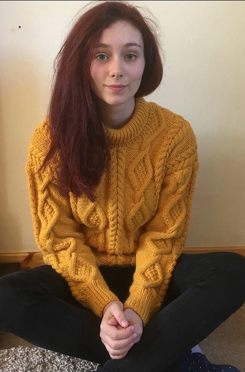 May include: A mustard yellow cable-knit jumper with a crew neck and diamond pattern on the sleeves and front. The jumper is worn by a person with long auburn hair and black trousers. The person is sitting cross-legged.