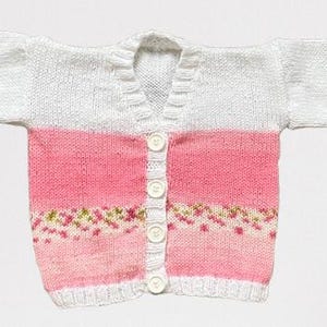 May include: A long-sleeved knit cardigan featuring a white upper section and a pink lower section. The garment has a V-neckline, a vertical row of four white buttons down the centre, and a decorative band of small green and pink floral patterns.
