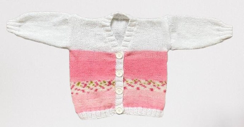 May include: A long-sleeved knit cardigan featuring a white upper section and a pink lower section. The garment has a V-neckline, a vertical row of four white buttons down the centre, and a decorative band of small green and pink floral patterns.