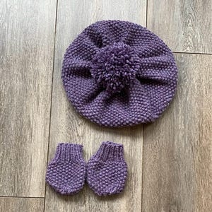 May include: A knitted purple baby hat with a pom-pom and matching mittens. The set is made of a textured yarn and is displayed on a wooden surface. The hat is round and the mittens are small.