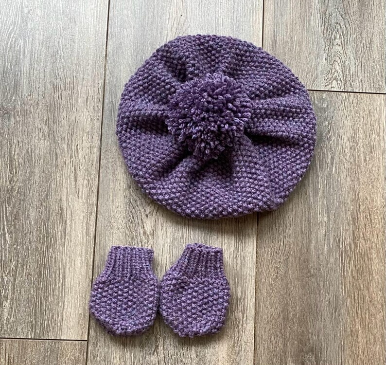 May include: A knitted purple baby hat with a pom-pom and matching mittens. The set is made of a textured yarn and is displayed on a wooden surface. The hat is round and the mittens are small.