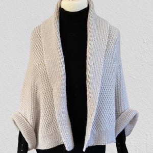 Op de afbeelding: Een beige gebreid vestje met een gestructureerd honingraatpatroon en een brede geribbelde sjaalkraag. Het kledingstuk heeft korte mouwen met omslag en wordt gedragen over een zwart shirt met lange mouwen en een col met zichtbare knopen.