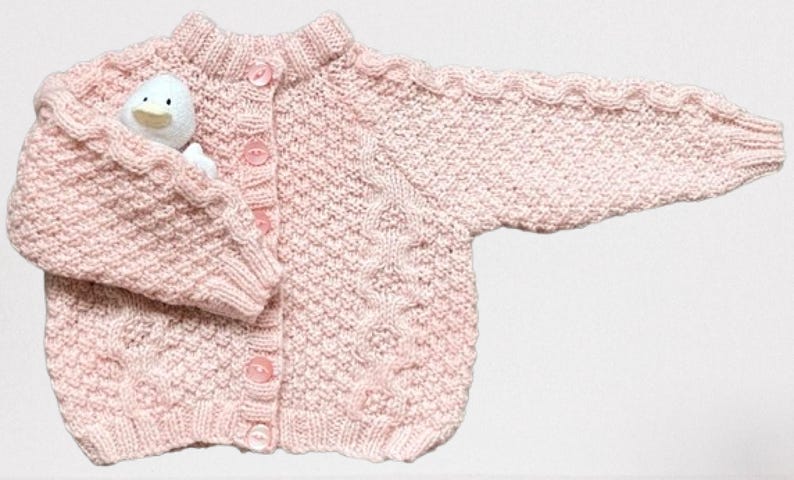 Op de afbeelding: Een lichtroze gebreid babyvestje met een knoopsluiting aan de voorkant en een klein wit stoffen eendje in de zak. Het vestje heeft een textuur gebreid patroon en is gemaakt van zacht garen.