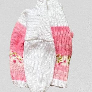 May include: A hand-knitted jumper featuring a white upper section and a pink lower section with a horizontal band of small, multi-coloured floral patterns. The garment has long sleeves, a ribbed neckline, and ribbed cuffs in a solid white colour.