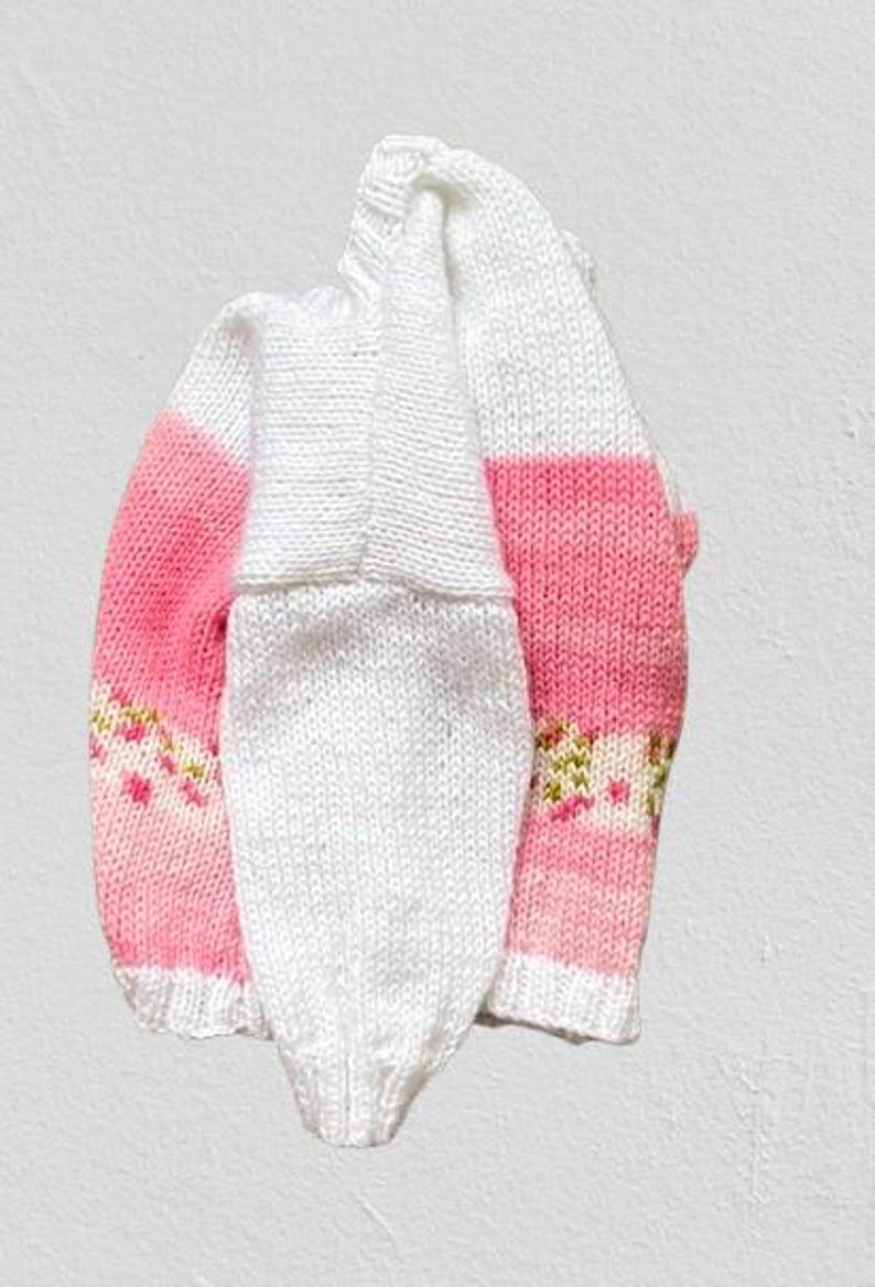 May include: A hand-knitted jumper featuring a white upper section and a pink lower section with a horizontal band of small, multi-coloured floral patterns. The garment has long sleeves, a ribbed neckline, and ribbed cuffs in a solid white colour.