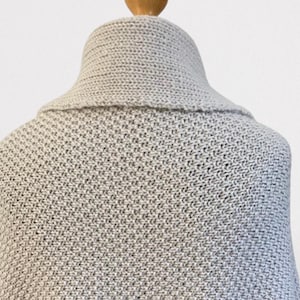 Op de afbeelding: Een lichtgrijze gebreide sjaal met een gestructureerd breipatroon en een brede, omgeslagen kraag. Het kledingstuk wordt getoond op een houten etalagepopnek, waarbij het dichte, uniforme weefsel van de stof en de vorm van de kraag zichtbaar zijn.