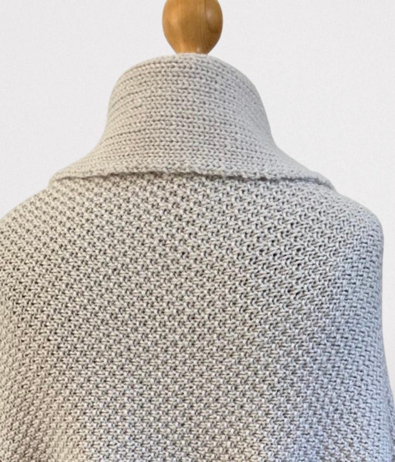 Op de afbeelding: Een lichtgrijze gebreide sjaal met een gestructureerd breipatroon en een brede, omgeslagen kraag. Het kledingstuk wordt getoond op een houten etalagepopnek, waarbij het dichte, uniforme weefsel van de stof en de vorm van de kraag zichtbaar zijn.