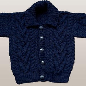 May include: A dark navy blue long-sleeve knit cardigan featuring a thick shawl collar and a vertical cable knit pattern. The front of the garment is secured with five round, metallic-toned buttons down the center, and the cuffs and hem are finished with a ribbed knit texture.