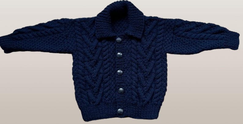 May include: A dark navy blue long-sleeve knit cardigan featuring a thick shawl collar and a vertical cable knit pattern. The front of the garment is secured with five round, metallic-toned buttons down the center, and the cuffs and hem are finished with a ribbed knit texture.
